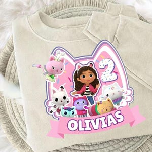 May include: A beige sweatshirt with a colourful graphic of a girl and cartoon cats. The design includes the number 2 and the name "OLIVIAS" on a pink banner, set against a pink cat-ear shaped background.