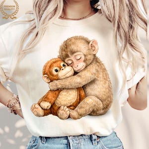 May include: Cream-colored t-shirt featuring a heartwarming illustration of two hugging monkeys. One monkey is brown, the other is orange. The image has a 'Premium Quality' seal in the upper left corner.