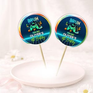 Può includere: Due lecca lecca rotondi a tema gaming. Ogni lecca lecca ha uno sfondo blu con una grafica di controller e il testo "BRUH", "YOU IN? OLIVER'S 10th Birthday". I lecca lecca sono su bastoncini di legno e poggiano su un piatto bianco.