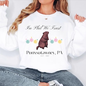 Può includere: Felpa bianca con un'illustrazione di marmotta marrone e il testo "In Phil We Trust" e "Punxsutawney, PA EST. 1887". La marmotta è circondata da fiori rosa, gialli e blu.