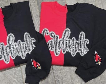 Puede incluir: Sudaderas y camisetas de manga larga rojas y negras. La palabra "Cardinals" está escrita en una fuente cursiva brillante en la parte delantera. Un logotipo de cardenal rojo está en la manga izquierda de la camiseta de manga larga.