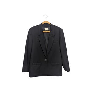 May include: A solid black blazer featuring a single-button closure, notched lapels, long sleeves, and two front flap pockets. The garment is displayed on a wooden hanger, showcasing a structured silhouette with a clean, minimalist design aesthetic.