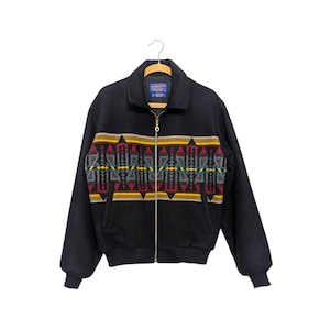 May include: A black zip-up jacket featuring a horizontal band across the chest with a geometric pattern in red, yellow, grey, and teal. The jacket has a collar, long sleeves with ribbed cuffs, a ribbed hem, and a Pendleton brand label at the neck.