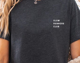 May include: A dark gray t-shirt with the text "Slow Runners Club" printed on the front. The shirt is being worn by a person with long blonde hair and is paired with denim shorts.