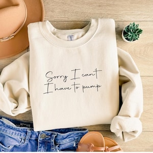 Può includere: Felpa color crema con la scritta "Sorry I can't I have to pump" in corsivo nero. La felpa è piegata su una superficie di legno, con jeans blu, sandali e un cappello sullo sfondo. È visibile anche una piccola pianta grassa in vaso.