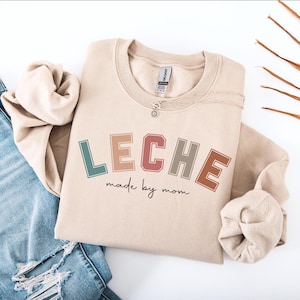 May include: A beige sweatshirt with the text "LECHE made by mom" in colorful letters. The letters are in a retro style with a rainbow color scheme.