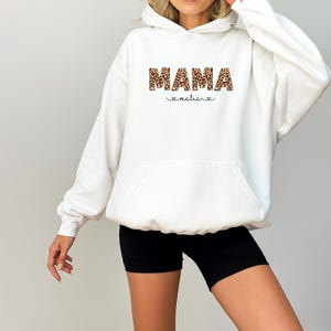 May include: A white hooded sweatshirt with a front pocket and a drawstring. The word "MAMA" is printed across the chest in a leopard print pattern, with the name "malia" below it in a script font, with two hearts.