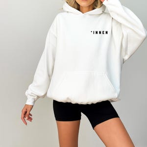 May include: White hoodie with a front pocket and the word "INNEN" printed in black on the upper chest. The hoodie is made of a soft material and has a drawstring hood. The model is wearing black shorts.