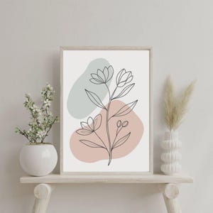 Puede incluir: Una impresión artística floral enmarcada con un contorno negro de una flor. El fondo tiene formas suaves en verde apagado y melocotón. La obra de arte se exhibe en una estantería de madera clara con jarrones decorativos.