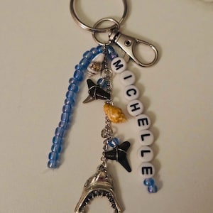 May include: A silver keychain with a lobster clasp and a split ring. The keychain features a blue beaded strand, a strand of white letter beads spelling "MICHELLE", and several shark-themed charms, including a shark jaw and shark teeth.