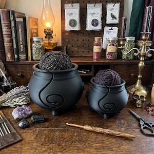 May include: Two matte black cauldron-shaped yarn bowls with spiral cutouts, each holding a ball of variegated yarn. The bowls sit on a wooden desk alongside a wooden crochet hook, metal knitting needles in a leather case, scissors, and crystals.