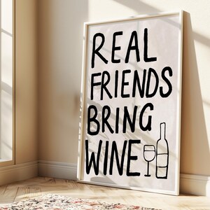 以下が含まれることがあります： 「REAL FRIENDS BRING WINE」というテキストが黒い太字で書かれたフレーム入りのプリント。テキストの下には、ワインボトルとワイングラスの黒いイラストがあります。プリントは白いフレームに入っており、壁に立てかけられています。