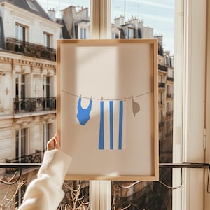 Peut inclure: Une affiche encadrée présentant une illustration minimaliste d'articles de plage. Un maillot de bain bleu, une serviette rayée et un chapeau de soleil sont suspendus à une corde à linge. L'œuvre est encadrée dans un cadre en bois.