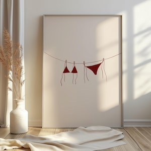 May include: Framed art print depicting a minimalist illustration of a red bikini set hanging on a washing line. The artwork is set against a neutral background, creating a clean and modern aesthetic. The print is displayed in a room with natural light.