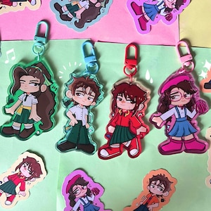 Puede incluir: Una colección de llaveros de acrílico con personajes ilustrados al estilo anime con diversos atuendos, incluyendo faldas, camisas y sombreros. Cada llavero tiene un cierre de plástico de color y muestra un diseño de personaje único.