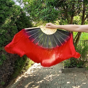 Puede incluir: Un abanico de seda rojo y negro, abierto sobre un fondo de follaje verde. El abanico tiene un marco de madera y un diseño calado decorativo en la parte superior. La parte roja es una tela fluida y semitransparente.