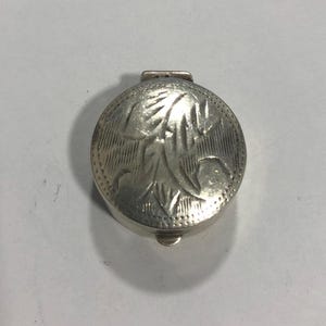 Puede incluir: Pequeño pastillero redondo plateado con tapa abisagrada. La tapa está decorada con un diseño floral en relieve y líneas verticales. El pastillero está cerrado y descansa sobre una superficie blanca.