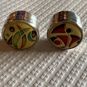 Può includere: Due gemelli argentati con disegni astratti colorati. Un gemello presenta uno sfondo dorato con forme verdi, marroni e turchesi. L'altro ha uno sfondo dorato con forme rosse, nere e bianche. I gemelli sono circolari e hanno una finitura lucida.