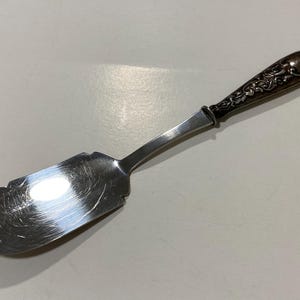 Op de afbeelding: Zilveren taartschep met een decoratief handvat. De schep heeft een breed, plat lemmet met een afgeschuinde rand. Het handvat heeft sierlijke bloemendetails en een donkere, mogelijk geoxideerde afwerking.