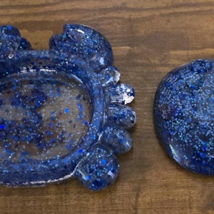 May include: Two resin ashtrays with a blue glitter design. One ashtray is shaped like a crab, and the other is a round, flat shape. Both are on a wooden surface.