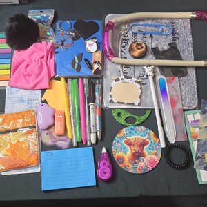 May include: A collection of stationery and desk accessories, including pens, sticky notes, a pink pouch, a blue notebook, and a desk lamp. There are also decorative items like stickers, a coaster with a cow, and a hair tie. The items are arranged on a dark surface.