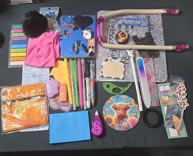 May include: A collection of stationery and desk accessories, including pens, sticky notes, a pink pouch, a blue notebook, and a desk lamp. There are also decorative items like stickers, a coaster with a cow, and a hair tie. The items are arranged on a dark surface.