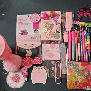 May include: A collection of pink stationery and beauty items, including a tumbler, pens, a notebook, face masks, and a sequined dolphin keychain. Other items include a pig-shaped sticky note, a floral print pouch, and a scrunchie.