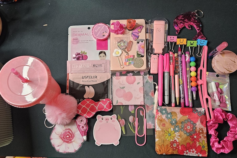 May include: A collection of pink stationery and beauty items, including a tumbler, pens, a notebook, face masks, and a sequined dolphin keychain. Other items include a pig-shaped sticky note, a floral print pouch, and a scrunchie.
