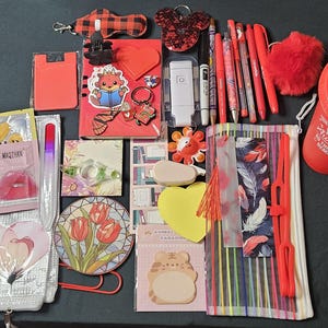 May include: A collection of red and silver stationery and accessories, including pens, a tumbler, a notebook, and a pencil case. There are also stickers, a nail file, and a heart-shaped paper clip. The items are arranged on a dark surface.