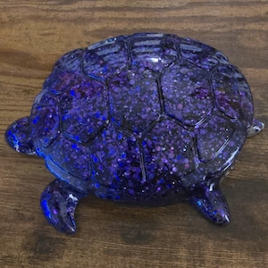 May include: A decorative turtle figurine with a glittery, iridescent finish. The turtle's shell is a dark blue with purple and silver glitter, and the body is a matching dark blue. The figurine has a glossy finish and is a unique home decor item.