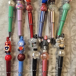 May include: A collection of decorative pens with various toppers. The pens feature designs such as a star, an egg with flowers, a heart with wings, a robot, a koala, a mouse, and a panda. The pens have different colored barrels, including green, pink, red, blue, and black.