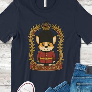 May include: Navy blue t-shirt featuring a cartoon corgi dressed as a Queen's Guard. The corgi is wearing a red uniform and a black hat, framed by a gold and red decorative border. The words "QUEEN'S GUARD" are printed below.