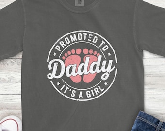 May include: A charcoal gray short-sleeve t-shirt featuring a circular white graphic with the text PROMOTED TO DADDY IT'S A GIRL and a pair of pink baby footprints in the center. The design has a distressed, weathered appearance.