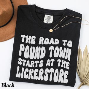 K&ouml;nnte beinhalten: Schwarzes T-Shirt mit dem wei&szlig;en Text "THE ROAD TO POUND TOWN STARTS AT THE LICKERSTORE", einer goldenen Halskette und einem braunen Hut. Das Shirt hat einen Rundhalsausschnitt und ein Comfort Colors-Etikett.