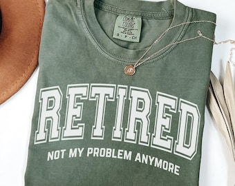 May include: A moss green short-sleeve t-shirt with a crew neckline. The shirt features the white text RETIRED NOT MY PROBLEM ANYMORE in a collegiate font. A Comfort Colors brand label is visible at the inner neck, and a gold necklace is present.