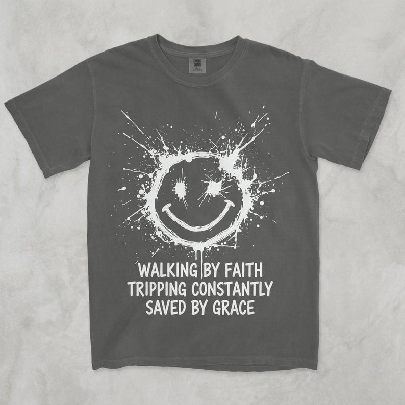 K&ouml;nnte beinhalten: Ein anthrazitfarbenes Kurzarm-T-Shirt mit einer wei&szlig;en Grafik eines farbspritzenden Smileys. Unter der Grafik steht der Text: WALKING BY FAITH, TRIPPING CONSTANTLY, SAVED BY GRACE. Das T-Shirt hat einen Rundhalsausschnitt und eine normale Passform.