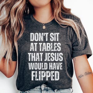 May include: A dark heather gray short-sleeve t-shirt featuring the white distressed text DON'T SIT AT TABLES THAT JESUS WOULD HAVE FLIPPED printed in a bold, sans-serif font across the center of the chest area of the garment.