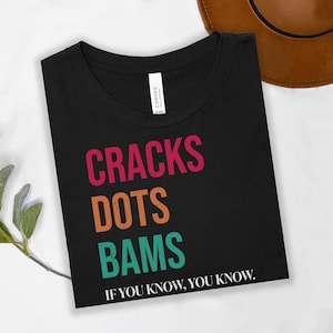 May include: Black t-shirt with the words "CRACKS DOTS BAMS" in pink, orange, and teal. The phrase "IF YOU KNOW, YOU KNOW" is printed below the words. A brown hat is partially visible in the background.