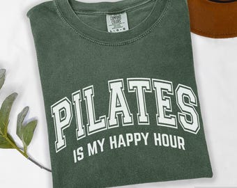 May include: A moss green crew neck t-shirt with a vintage-washed finish. The front features white collegiate-style lettering that reads PILATES IS MY HAPPY HOUR. The inner neck label displays the Comfort Colors brand logo and size L.