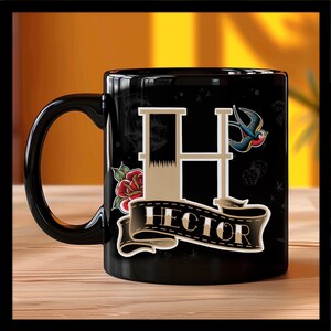 Puede incluir: Taza de cerámica negra con una letra blanca "H" y la palabra "HECTOR" en una pancarta. La taza tiene un diseño de estilo tatuaje con una rosa roja, un pájaro azul y dados.