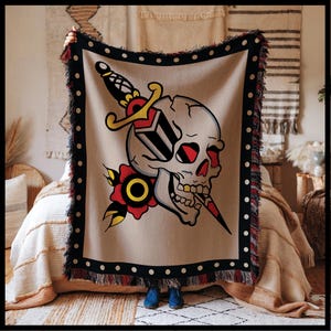 Op de afbeelding: Een geweven tapijtdeken met een traditioneel tatoeage-stijl ontwerp van een schedel en dolk. De schedel is wit met rode ogen en een gouden tand. De dolk is zwart en wit met een gouden handvat. De achtergrond is beige met zwarte stippen.