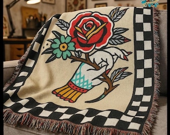 May include: Beige throw blanket featuring a hand holding a rose and a green flower. The blanket has a black and white checkered border and fringe. The artwork is in a tattoo style.