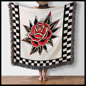 May include: A woven throw blanket with a red and black chequered border and a large red rose with green leaves and gold accents in the centre.