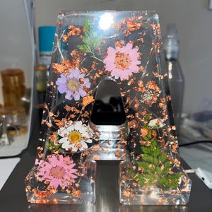 Puede incluir: Letra "A" de resina transparente con flores secas y copos de cobre incrustados. La letra presenta flores rosas, blancas y moradas, junto con follaje verde. La resina transparente permite ver los detalles.