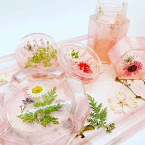 Peut inclure: Un ensemble de récipients en résine rose avec des fleurs et des feuilles pressées. Les récipients sont de formes et de tailles variées, notamment un bocal rond, un bocal carré et un plateau rectangulaire. Les fleurs et les feuilles sont pressées dans la résine, créant un design unique et magnifique.