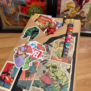 May include: A brown paper gift bag decorated with comic book characters, including Hulk, Thor, Iron Man, and the Avengers. The bag has a pencil with a superhero character design on it.