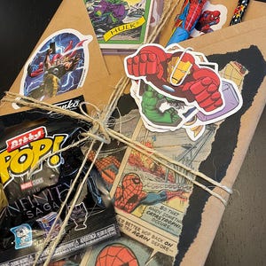 May include: A brown paper package wrapped with twine and decorated with various Marvel superhero stickers, including Hulk, Iron Man, and Spider-Man. The package also includes a small plastic bag with a Marvel Infinity Saga Pop figure inside.