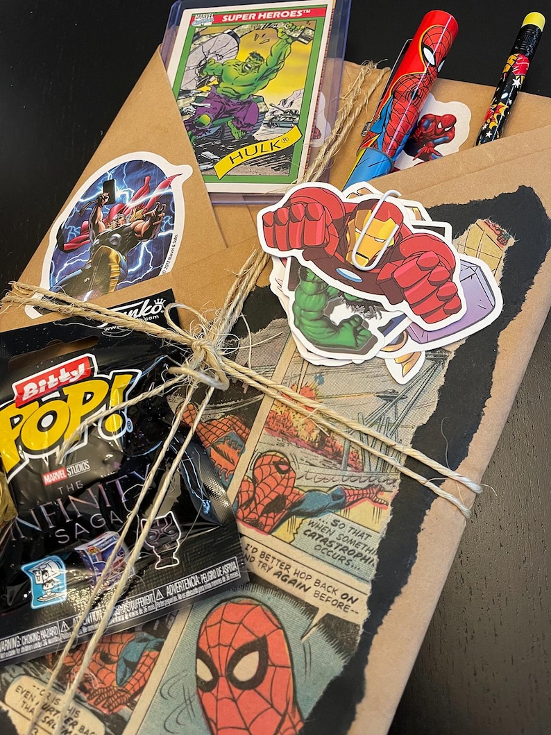May include: A brown paper package wrapped with twine and decorated with various Marvel superhero stickers, including Hulk, Iron Man, and Spider-Man. The package also includes a small plastic bag with a Marvel Infinity Saga Pop figure inside.