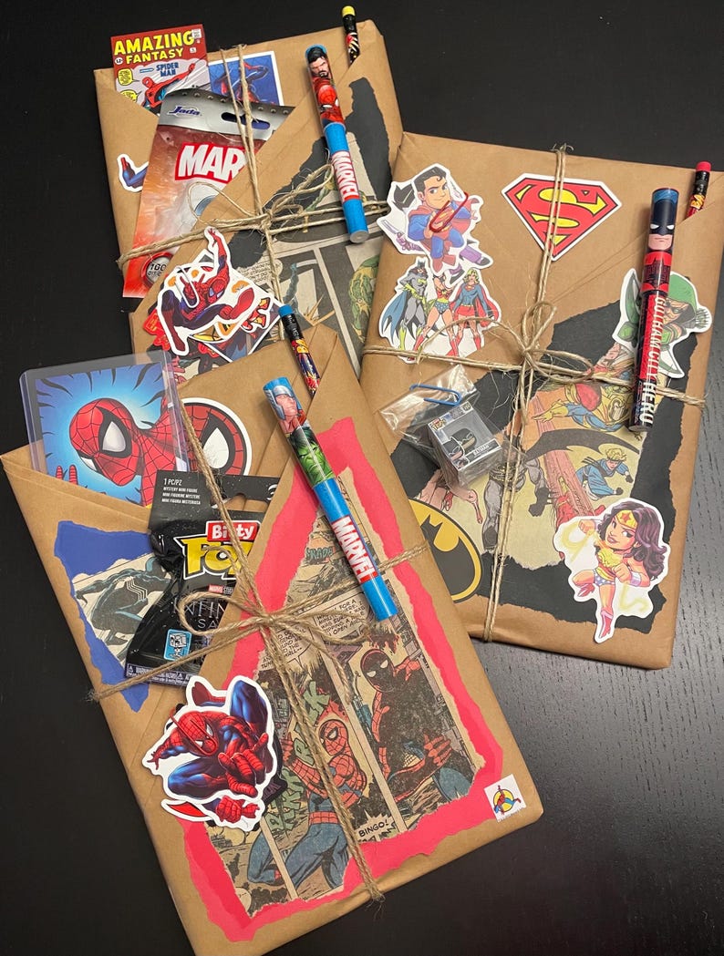 May include: Three brown paper packages wrapped with twine and decorated with superhero comic book covers and stickers. The packages include pencils with superhero logos.