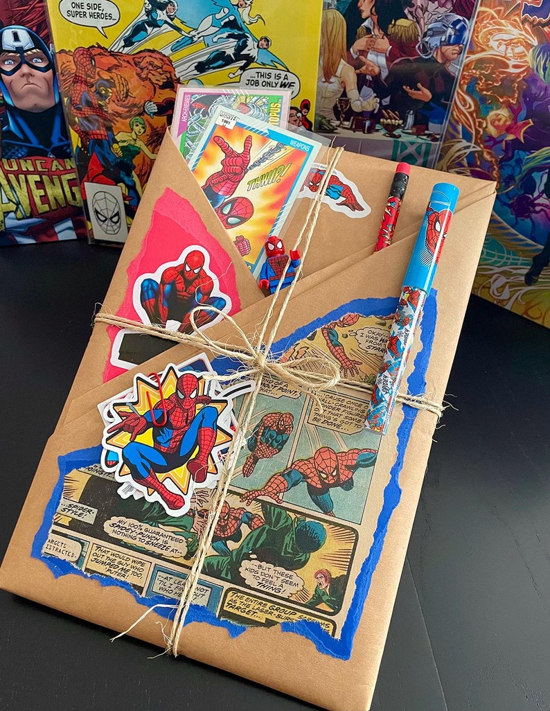 May include: A brown paper package wrapped with twine and decorated with blue and red comic book cutouts and stickers featuring Spiderman. The package includes a blue and red pen and a pencil.